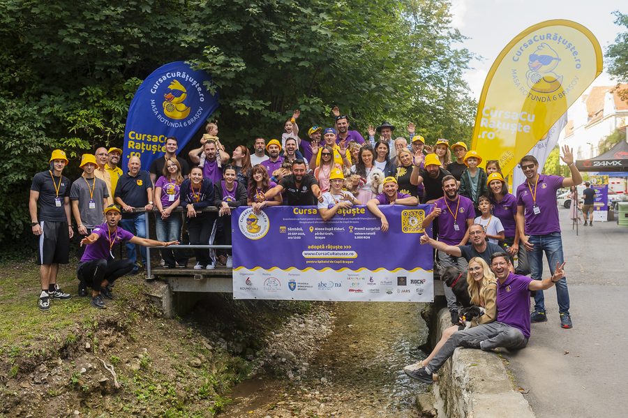 Echipa Cursei cu Rățuște 2025 pe pod, cu familiile, steaguri și bannerul cursei, cu mâinile ridicate.