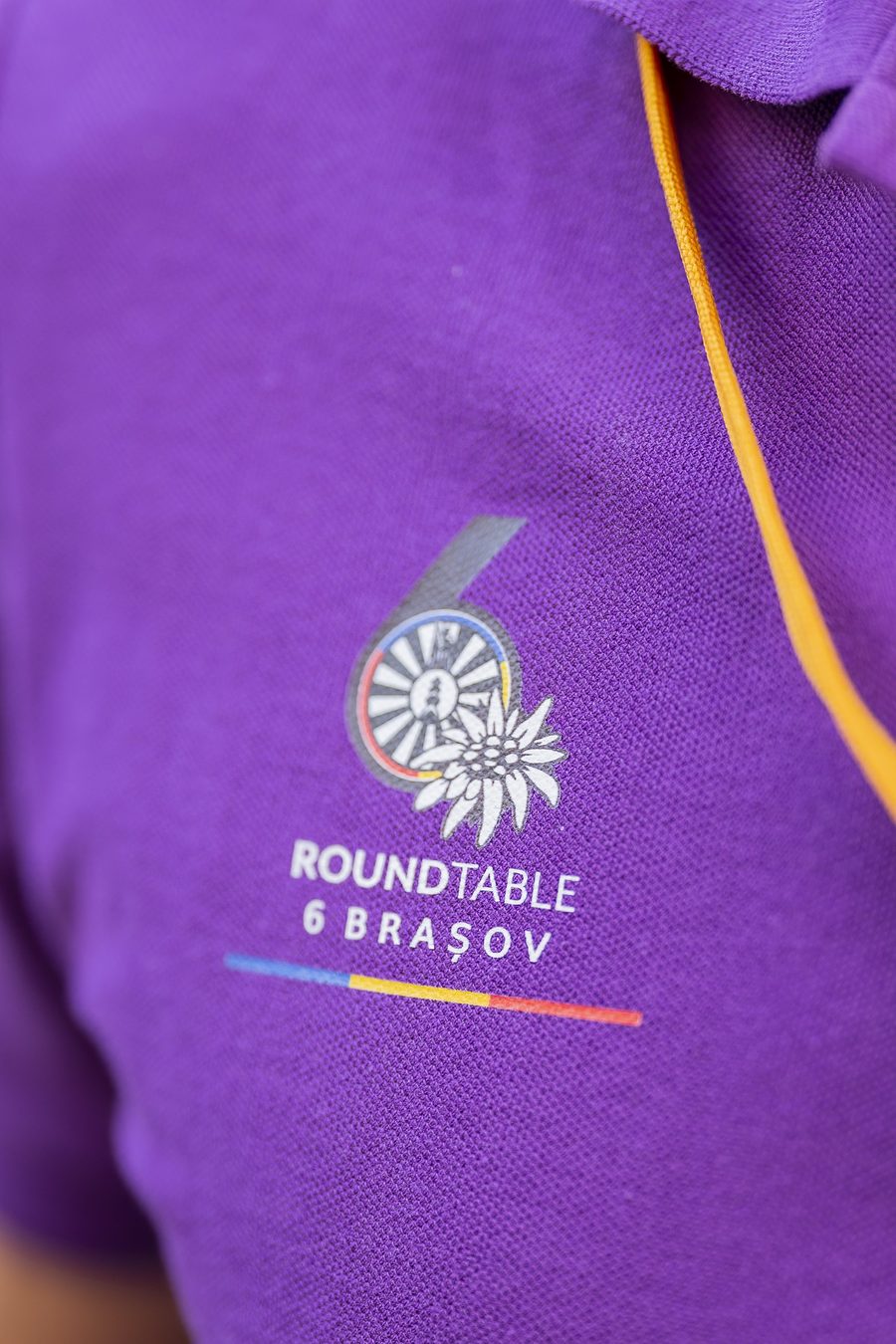 Emblema brodată pe un tricou mov: cifra 6, roata Round Table, o floare de colț și textul Roundtable 6 Brașov.